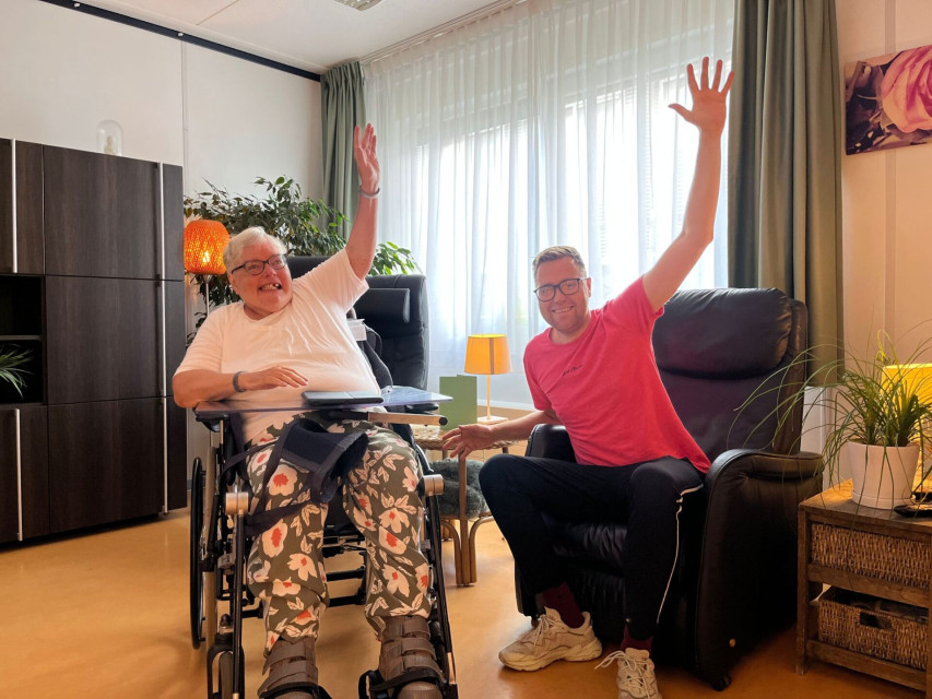Een zorgmedewerker juicht met een oudere vrouw in een rolstoel.